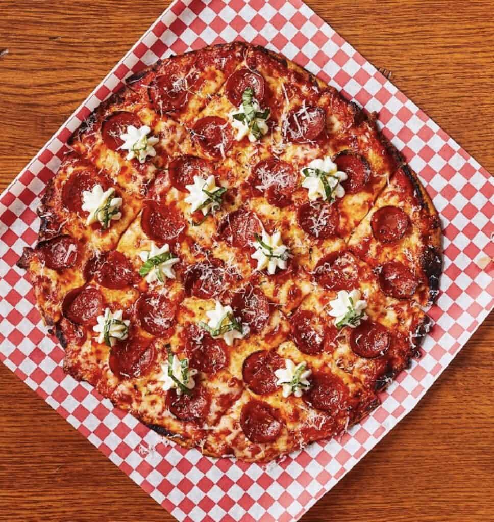 A pepperoni pizza on a red and white checkered paper, topped with dollops of ricotta cheese and grated parmesan, on a wooden table.