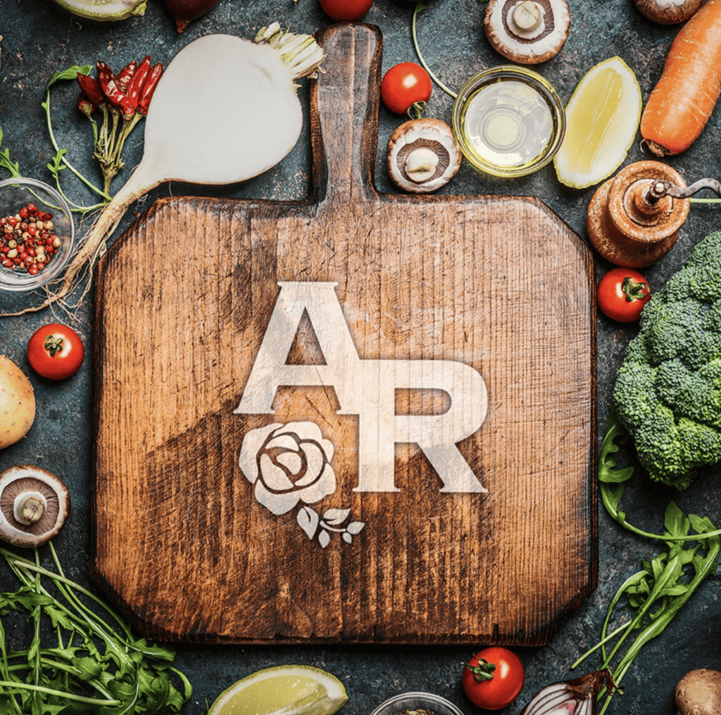 A wooden cutting board with "AR" and a rose engraved on it, surrounded by assorted fresh vegetables, herbs, and kitchen ingredients.