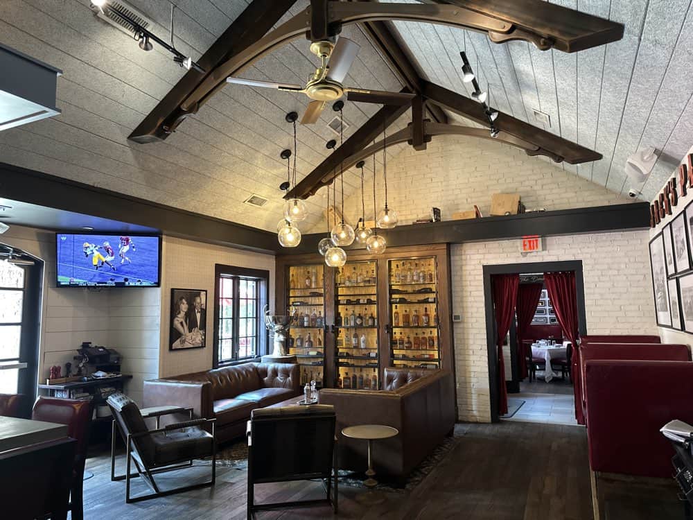 A cozy, rustic room with a high ceiling and exposed beams, featuring a leather seating area, a TV displaying a football game, shelves with bottles, and a doorway leading to another room.