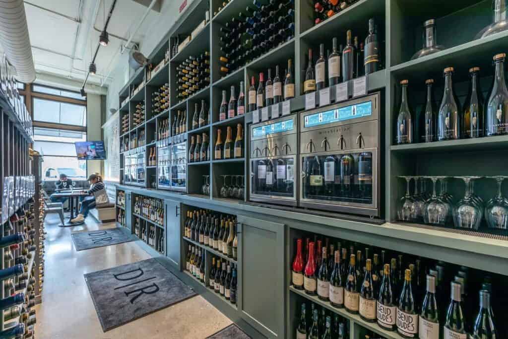Modern wine bar interior with shelves lined with bottles, wine dispensers, and glasses, with patrons seated near large windows in the background.