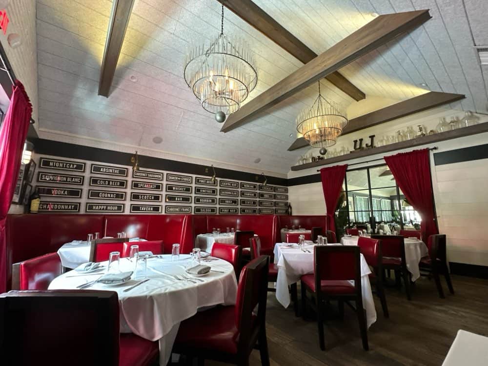 A restaurant interior featuring red booths, round tables with white tablecloths, chandeliers, and wine selections displayed on a wall-mounted menu.