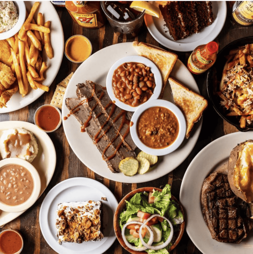 An overhead view of assorted barbecue dishes, including brisket, beans, toast, fries, salad, cake, and drinks, arranged on a wooden table.