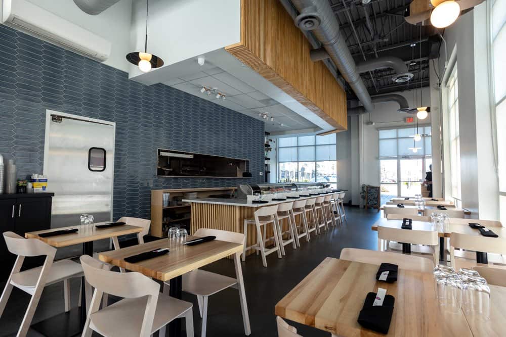 Modern cafe interior with wooden tables, white chairs, and a long counter. Large windows allow natural light, and industrial ceiling details are visible.