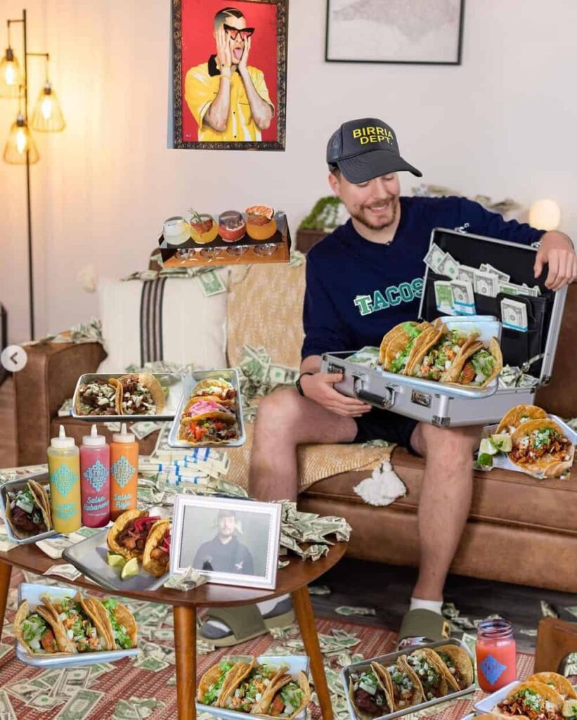 In a freestanding restaurant in downtown Duluth, a man seated on a couch holds a suitcase filled with tacos and cash. Around him are more tacos, hot sauce bottles, and a drink can. Walls feature playful art and photos, as if the place itself is infused with vibrant personality.