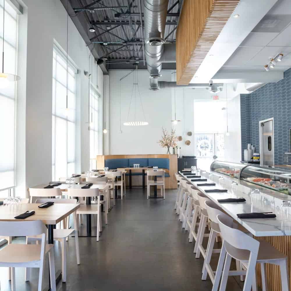 Modern restaurant interior with white chairs and tables, a long counter with bar stools, and large windows providing natural light. The ceiling features exposed ducts and lighting fixtures.