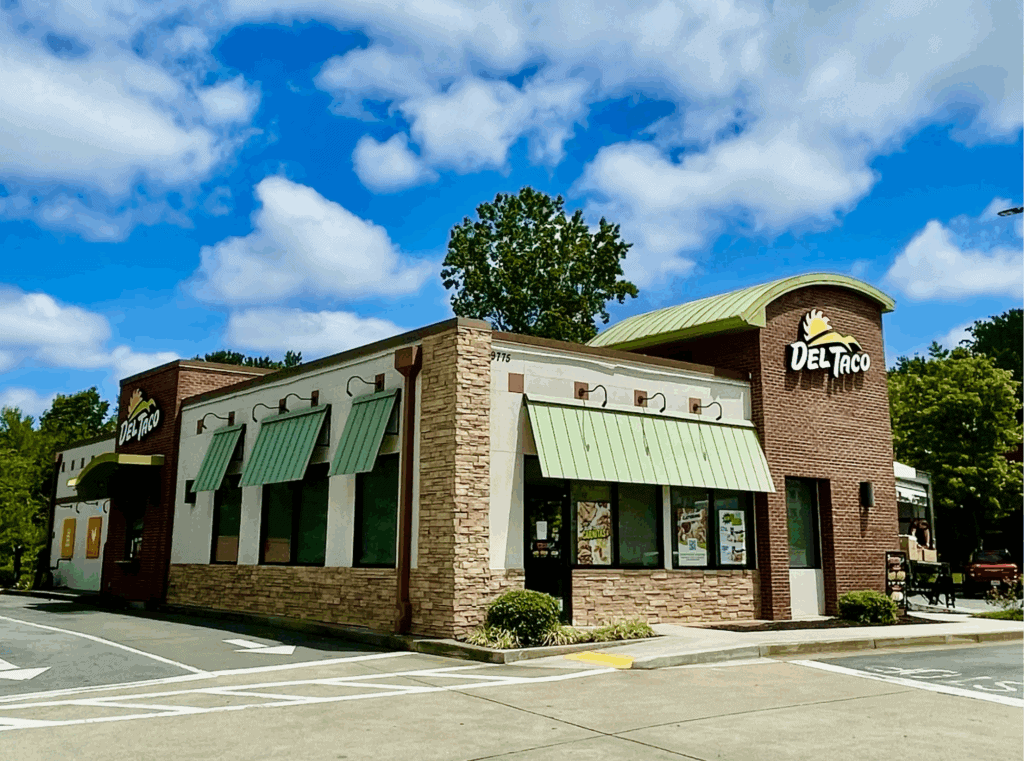 A Del Taco fast food restaurant in Metro Atlanta with green awnings, brick and stone exterior, and a drive-thru lane, set against a partly cloudy sky.