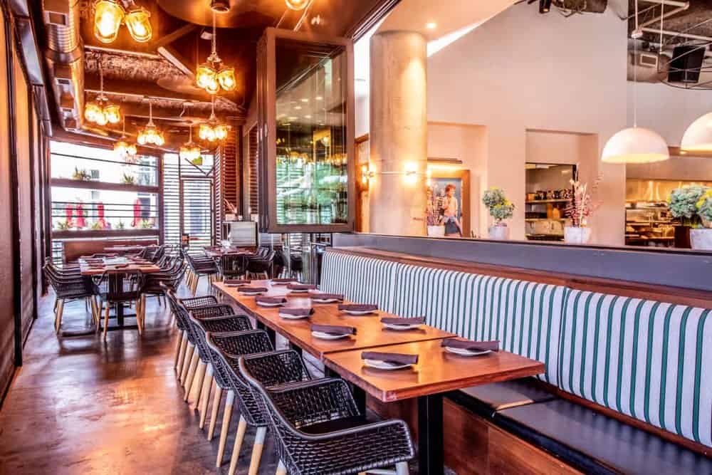 Modern restaurant interior with striped banquette seating, wooden tables set with plates, black chairs, pendant lights, and an open kitchen area in the background.