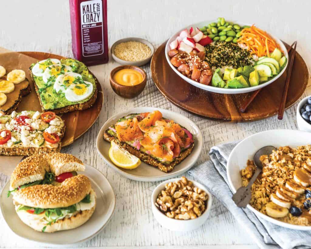 A variety of breakfast dishes including avocado toast, smoked salmon toast, a bagel sandwich, a grain bowl, granola with fruit, nuts, sauce, and a bottle of juice on a white table at this East Cobb GA Juice Franchise.