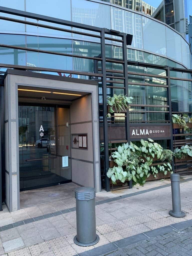 Street view of the entrance to ALMA Cocina, a modern restaurant with glass walls, metal framework, and large green plants by the doorway.