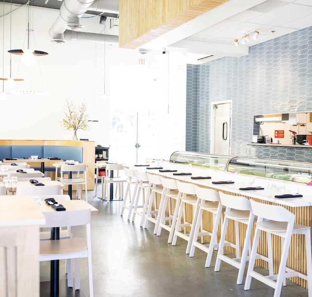 Bright, modern restaurant interior with white chairs, long counter, blue tile wall, and large windows. Tables are set with black napkins.